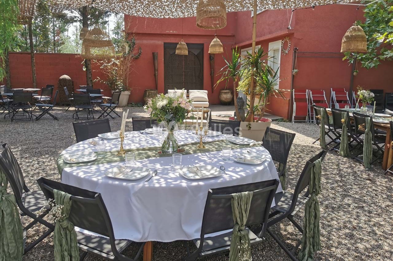 Table ronde dressée avec une nappe blanche, entourée de chaises décorées, dans une cour extérieure ombragée.