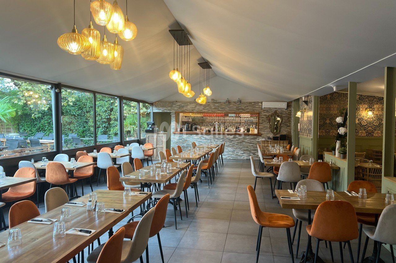Salle de restaurant lumineuse avec tables en bois, chaises colorées et bar en pierre au fond, éclairée par des suspensions…