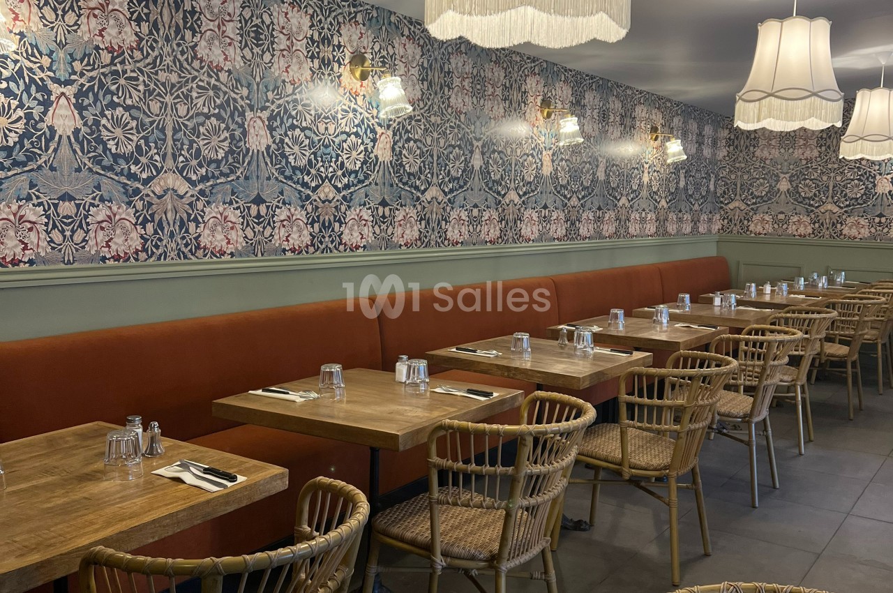 Salle de restaurant avec tables en bois, chaises en rotin, banquette orange et murs ornés d’un papier peint fleuri.