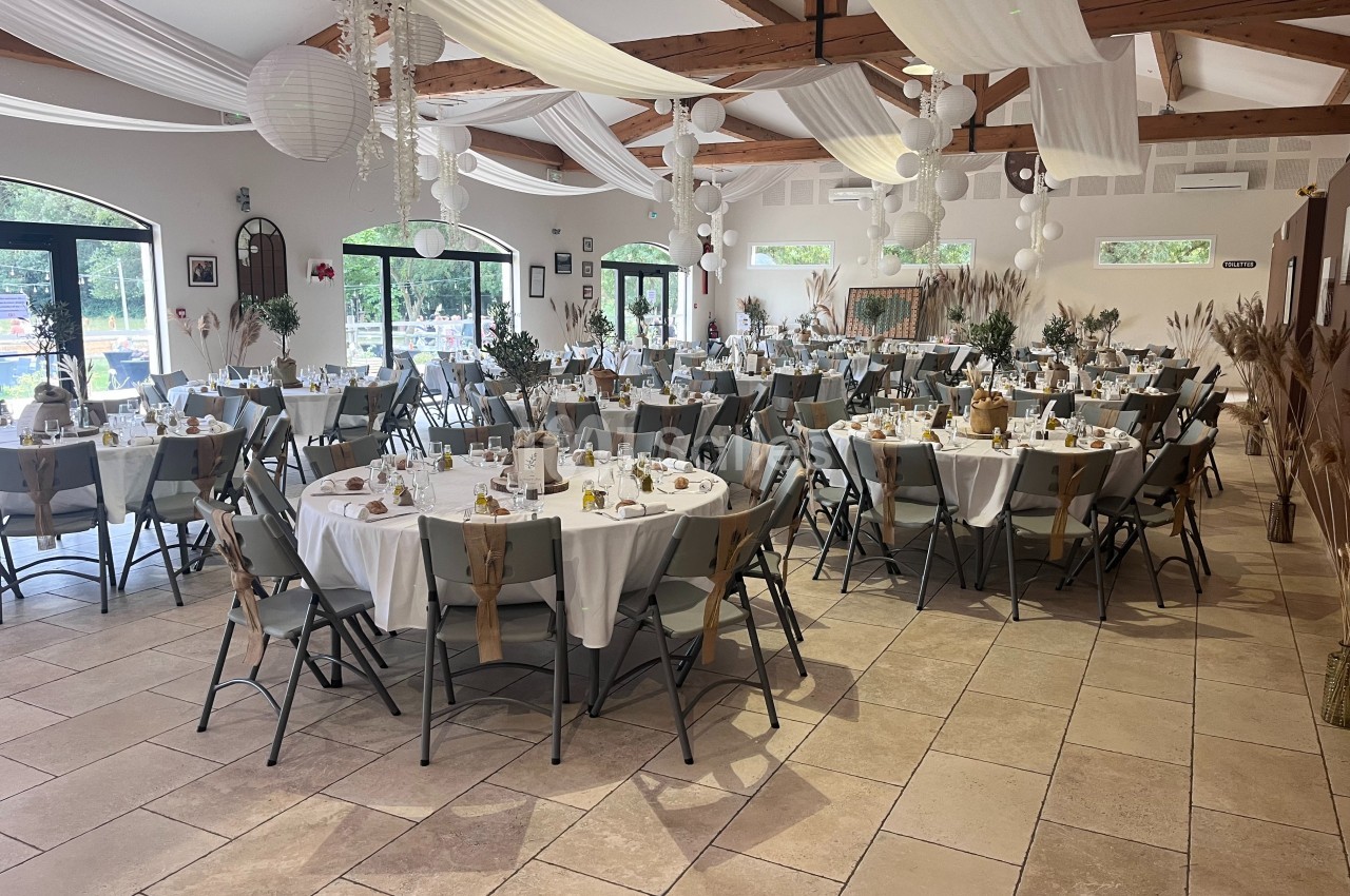 Salle de réception décorée pour un événement, avec tables rondes dressées, chaises pliantes et ornements suspendus.