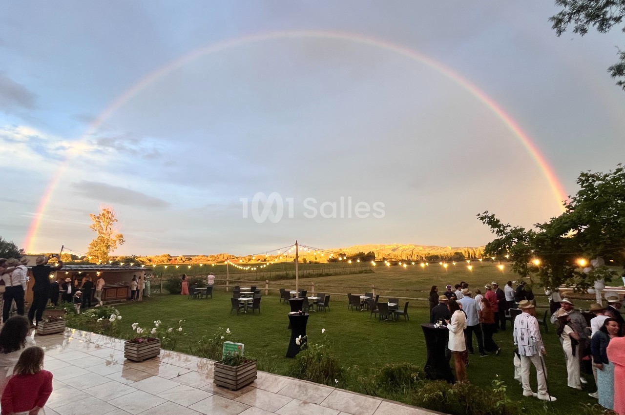 Un arc-en-ciel lumineux surplombe un jardin avec des invités rassemblés lors d'un événement en plein air.