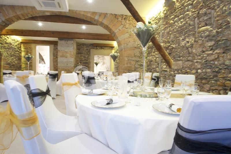 Salle de réception avec tables rondes dressées, nappes blanches et chaises décorées de rubans noirs et dorés.