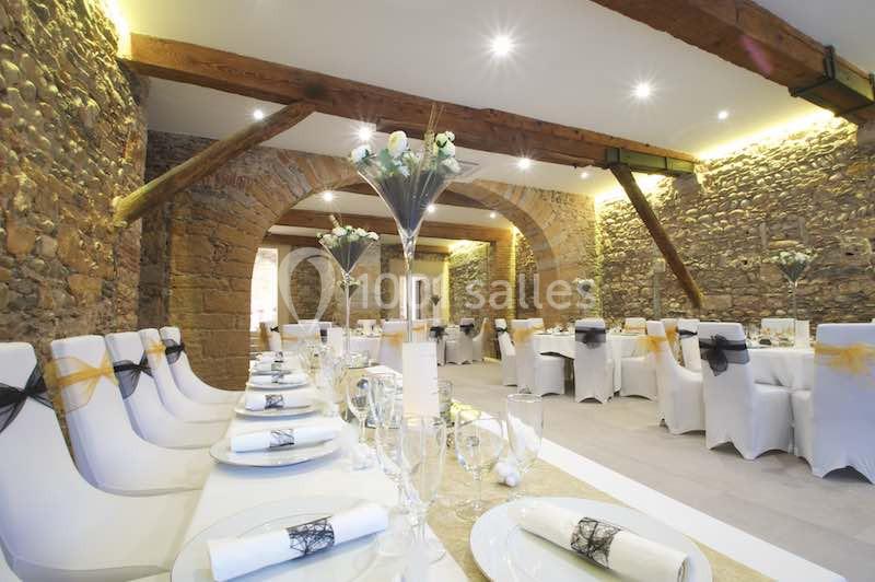 Salle de réception avec murs en pierre, poutres apparentes et tables décorées de nappes blanches et centres de table floraux.