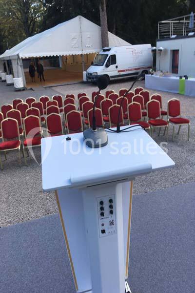 Un pupitre équipé de micros devant des rangées de chaises rouges disposées en extérieur, près d'une tente blanche.