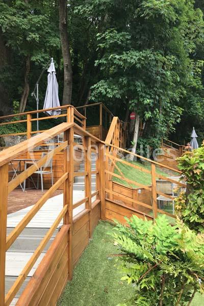 Terrasse en bois avec escaliers et rampes, entourée de végétation dense et de parasols fermés.