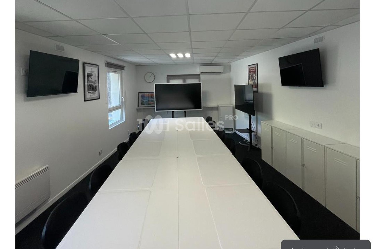 Salle de réunion lumineuse avec une grande table blanche, des chaises noires, plusieurs écrans et des affiches aux murs.