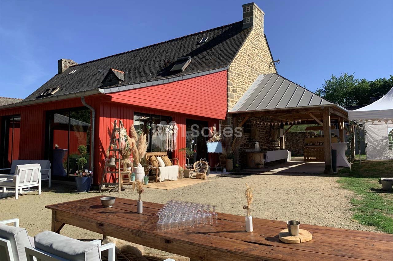 Maison en pierre avec extension rouge, terrasse aménagée, grande table en bois et espace couvert dans un jardin ensoleillé.