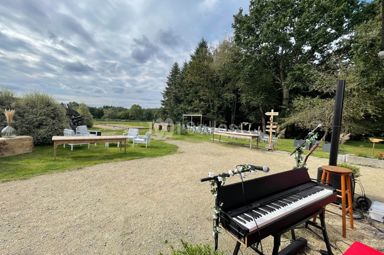 Piano décoré en extérieur sur une terrasse en gravier, entouré de mobilier de jardin et d'arbres sous un ciel nuageux.