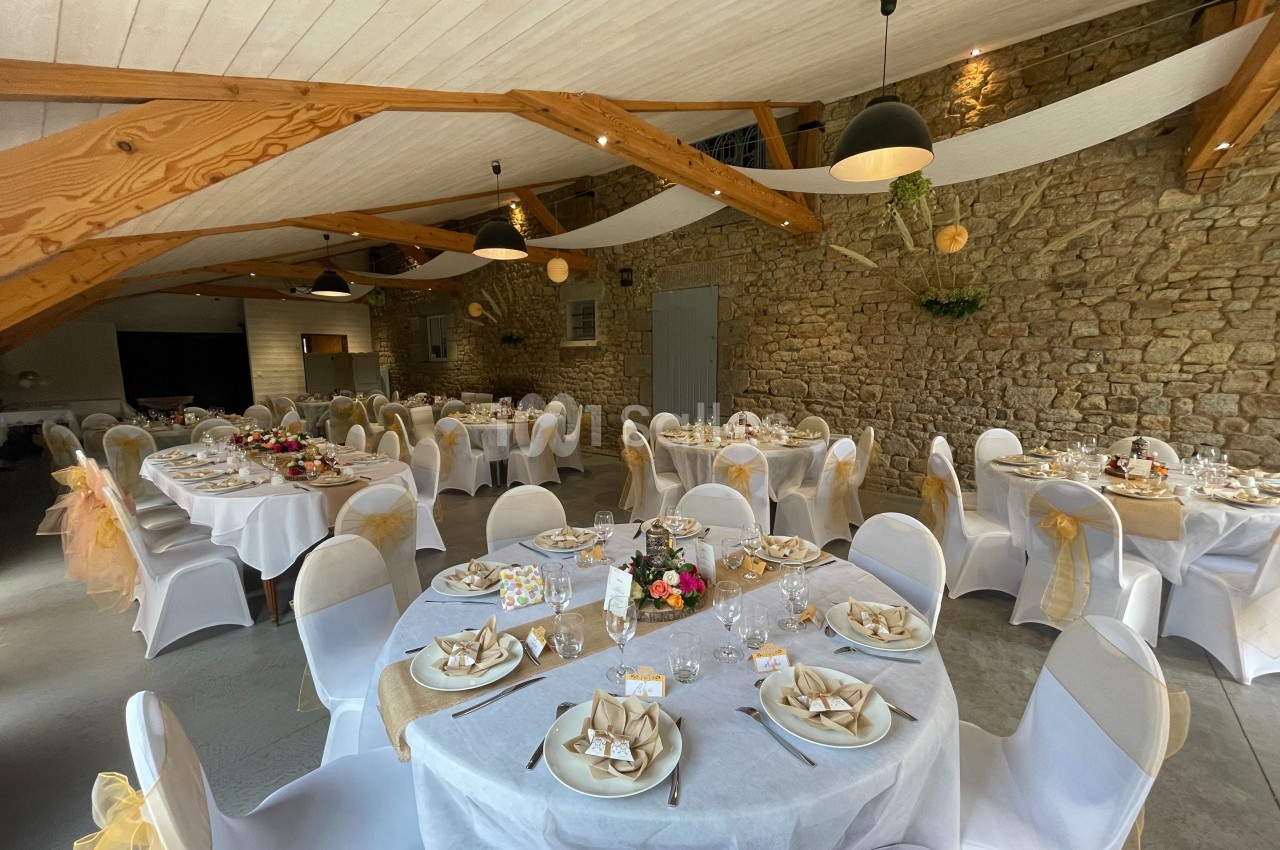 Salle de réception décorée avec des tables rondes dressées, nappes blanches, chaises ornées et murs en pierre apparente.