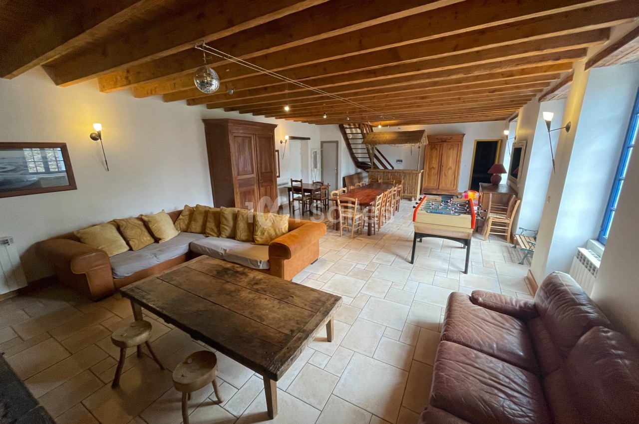 Salle de séjour spacieuse avec poutres apparentes, canapés, table en bois, baby-foot et grande table à manger.