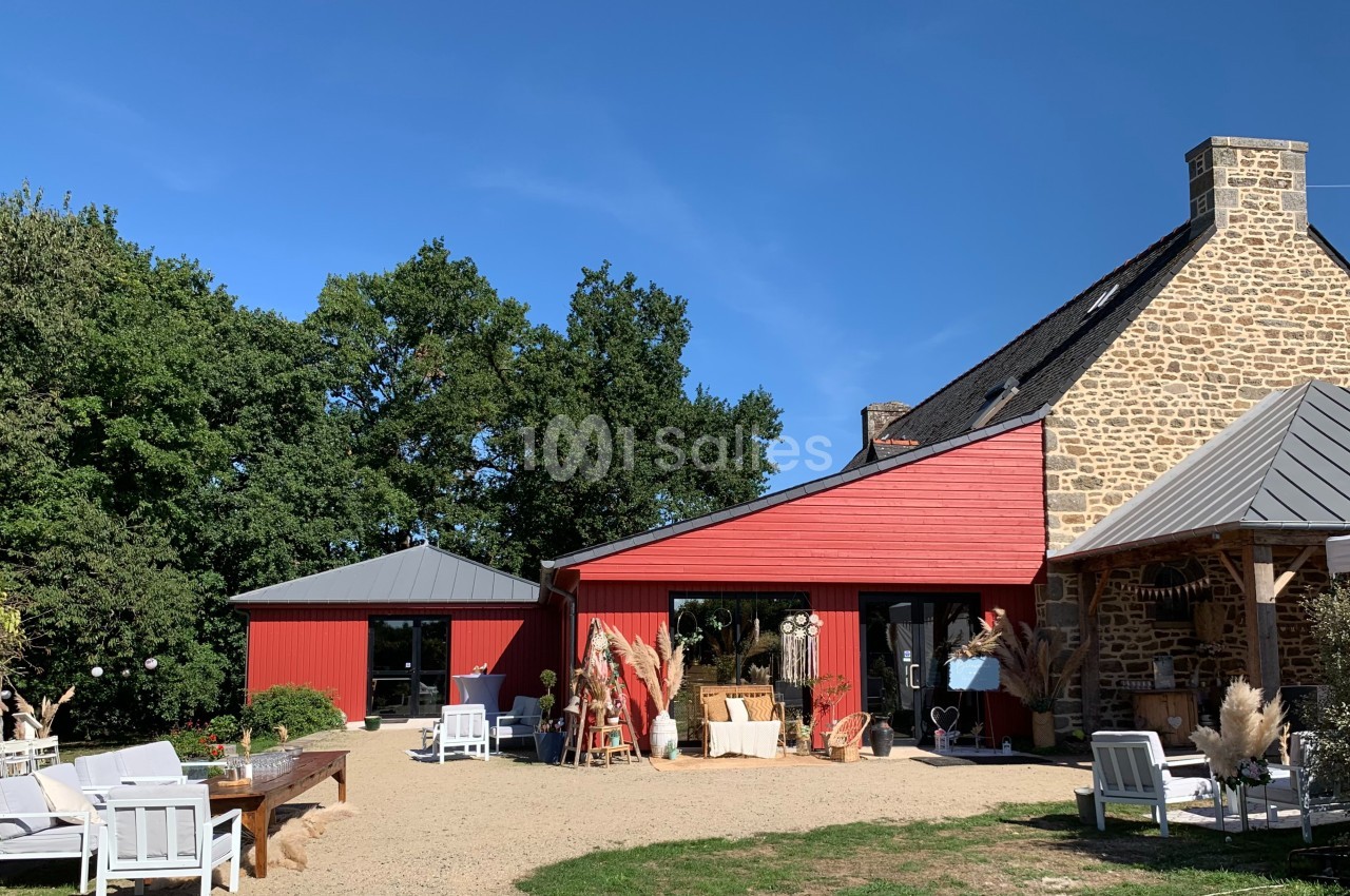 Bâtiment en pierre avec extension rouge, entouré de mobilier d'extérieur et de végétation sous un ciel bleu.