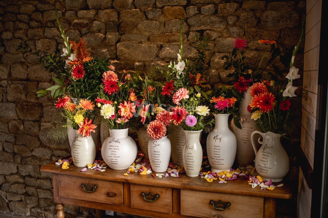 Vases en céramique blanche remplis de fleurs colorées, disposés sur une table en bois devant un mur en pierre.
