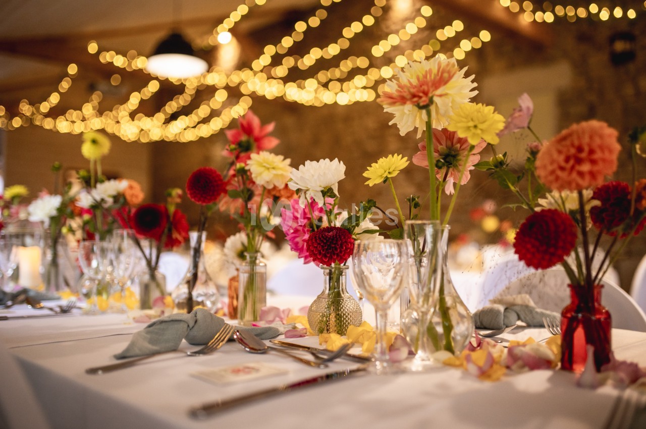 Tables décorées de bouquets colorés et de pétales, éclairées par des guirlandes lumineuses dans une salle chaleureuse.