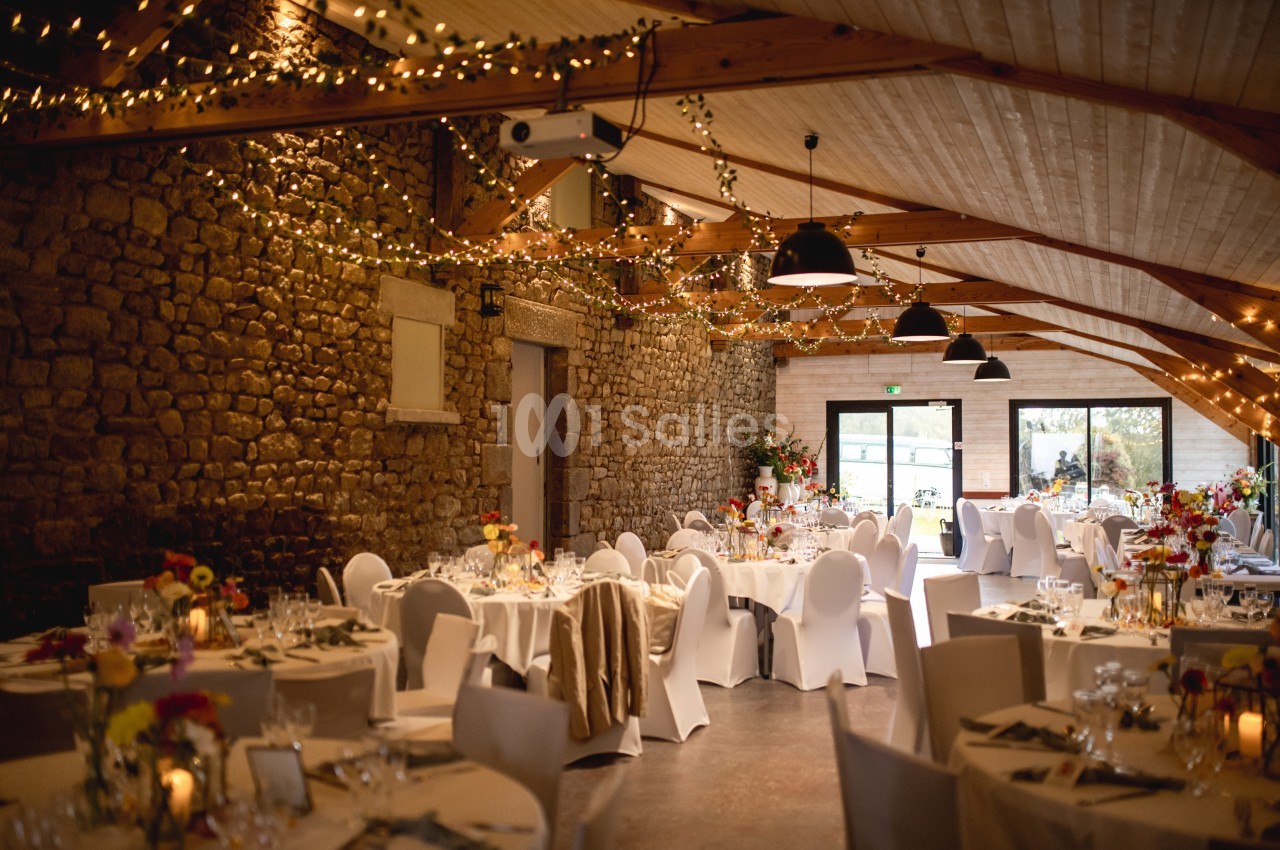 Salle de réception décorée avec des guirlandes lumineuses, des tables rondes dressées et des murs en pierre apparente.