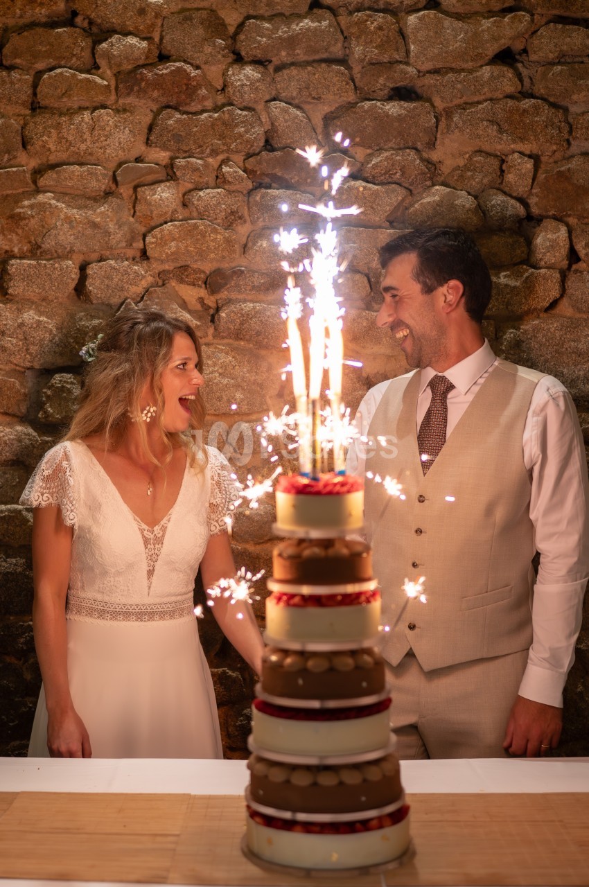 Un couple souriant devant une pièce montée décorée de cierges étincelants, sur fond de mur en pierre.