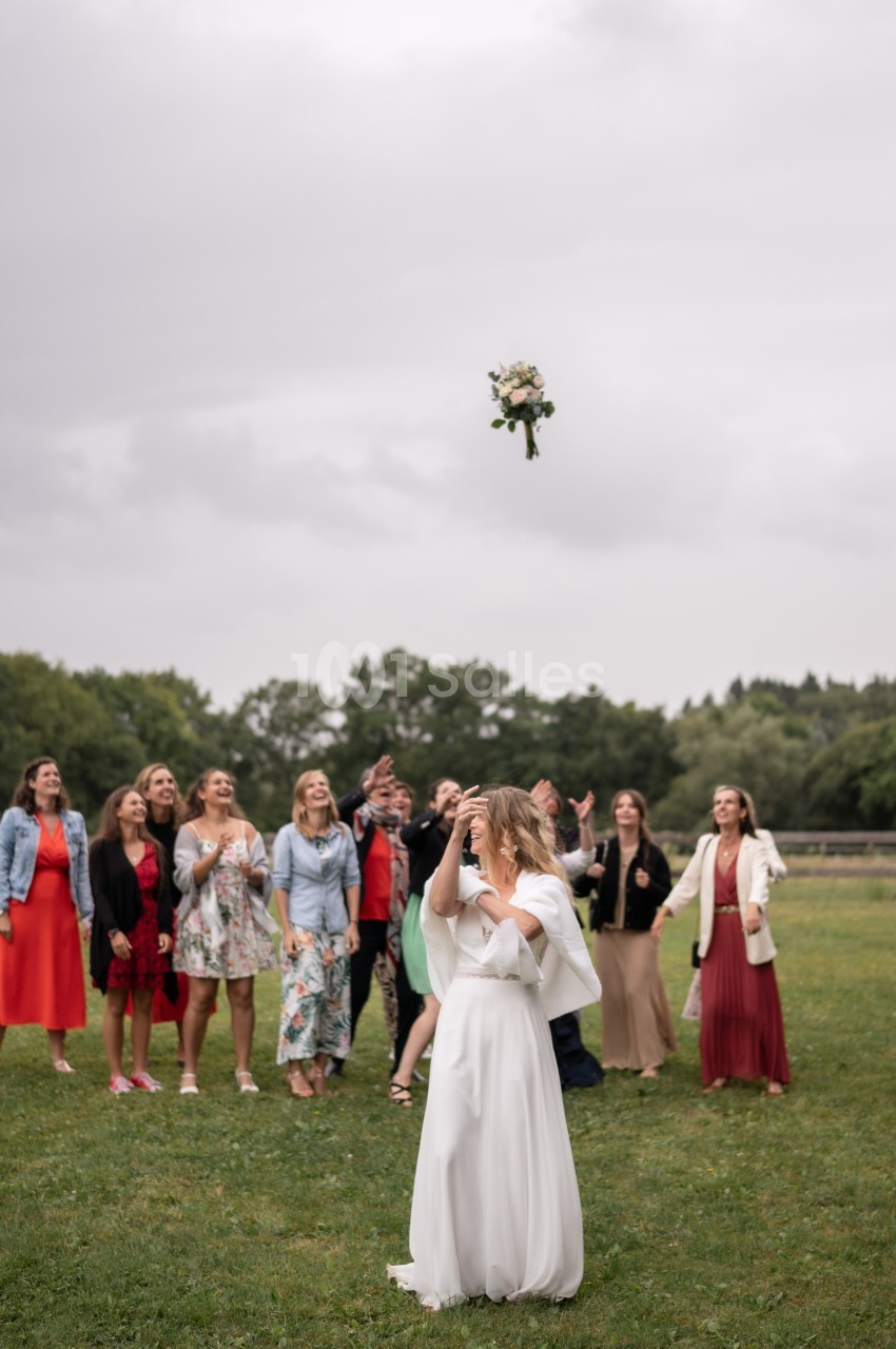 Une mariée lance son bouquet de fleurs vers un groupe de femmes dans un champ sous un ciel nuageux.