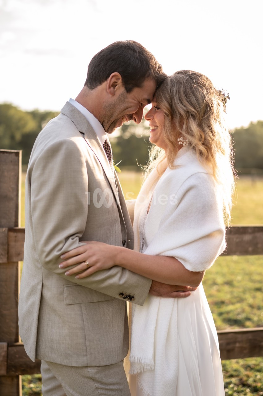 Un couple souriant s'enlace tendrement devant une clôture en bois, sous une lumière douce au coucher du soleil.