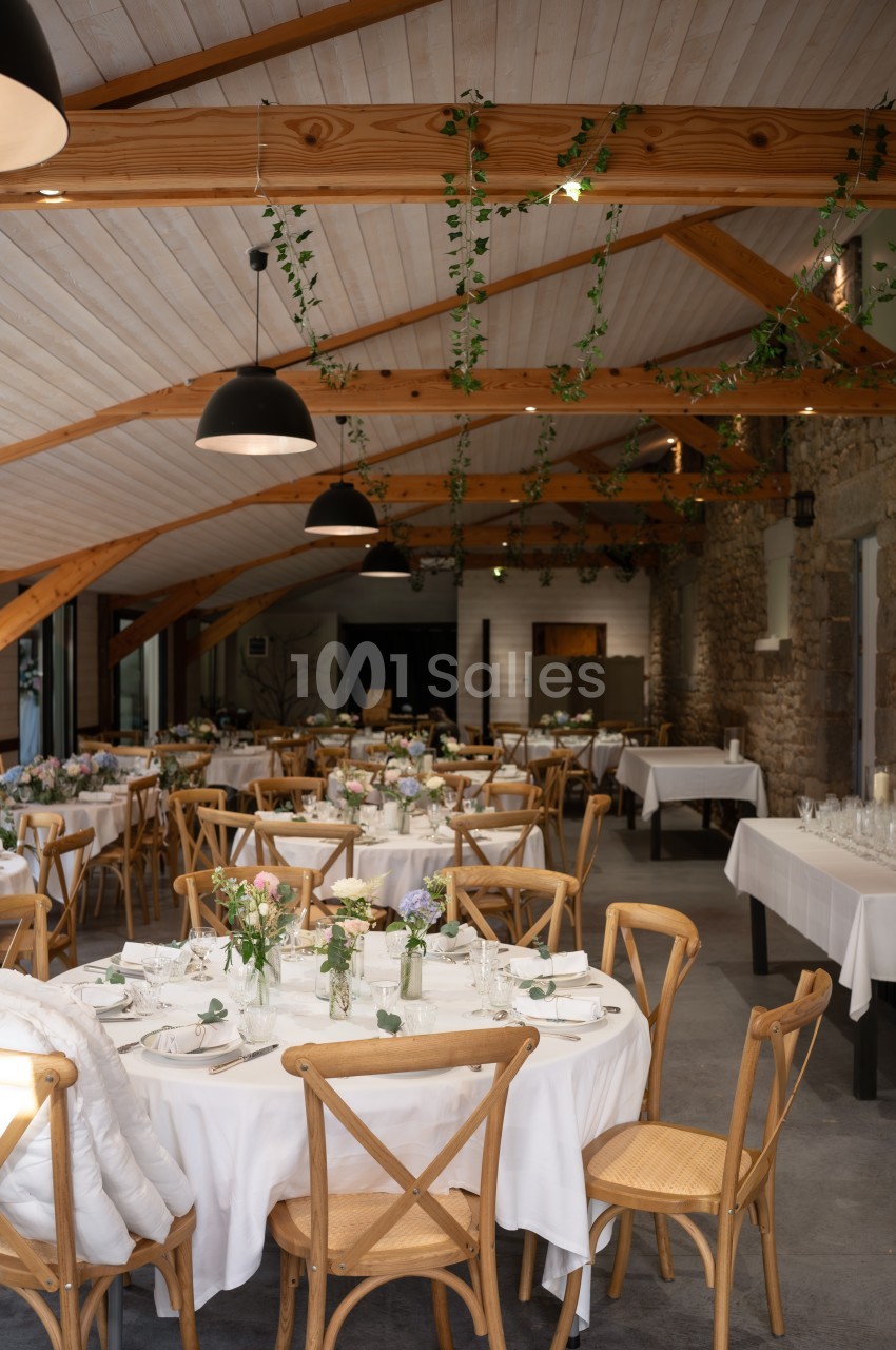 Salle de réception lumineuse avec tables rondes décorées de nappes blanches, fleurs et chaises en bois clair.