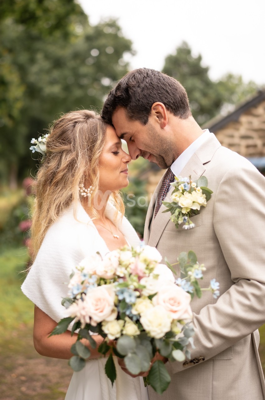 Un couple en tenue de mariage se tient face à face dans un jardin, tenant un bouquet de fleurs pastel.