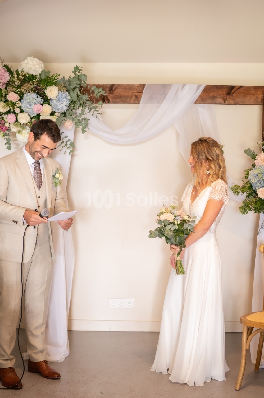 Un couple échange des vœux devant une arche décorée de fleurs lors d'une cérémonie de mariage en intérieur.