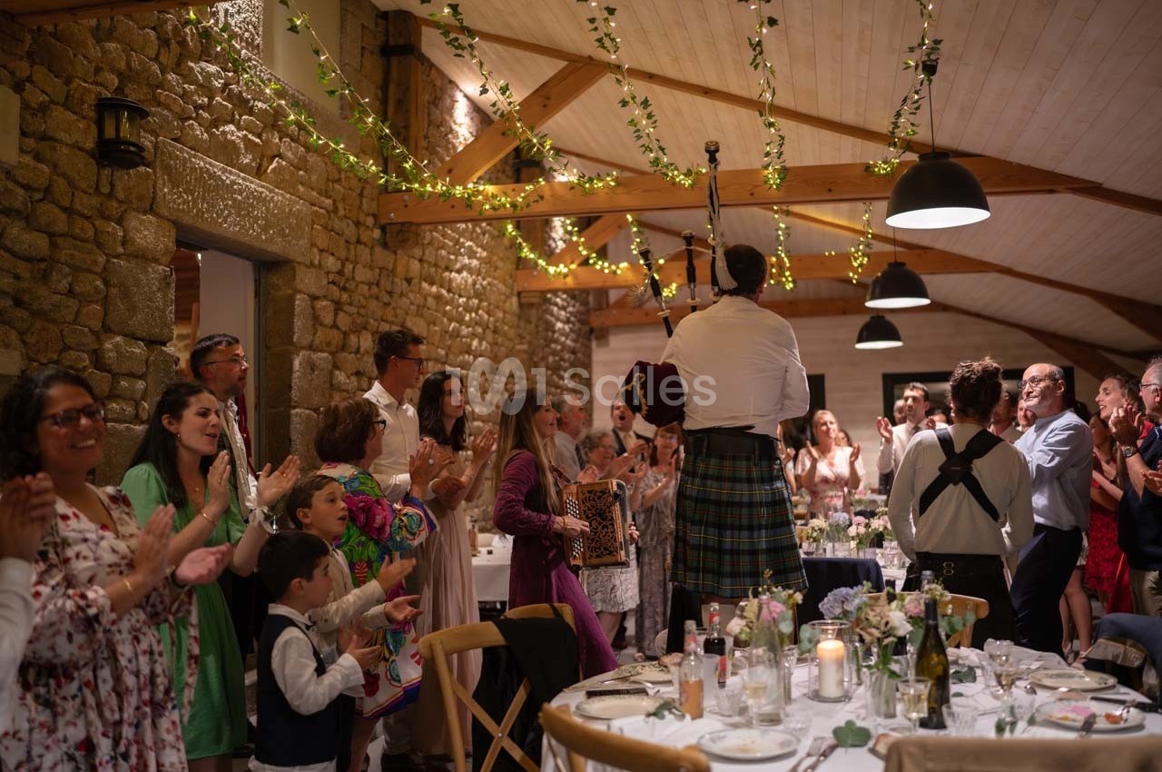 Un joueur de cornemuse en kilt joue devant des invités debout dans une salle de réception décorée de guirlandes lumineuses.