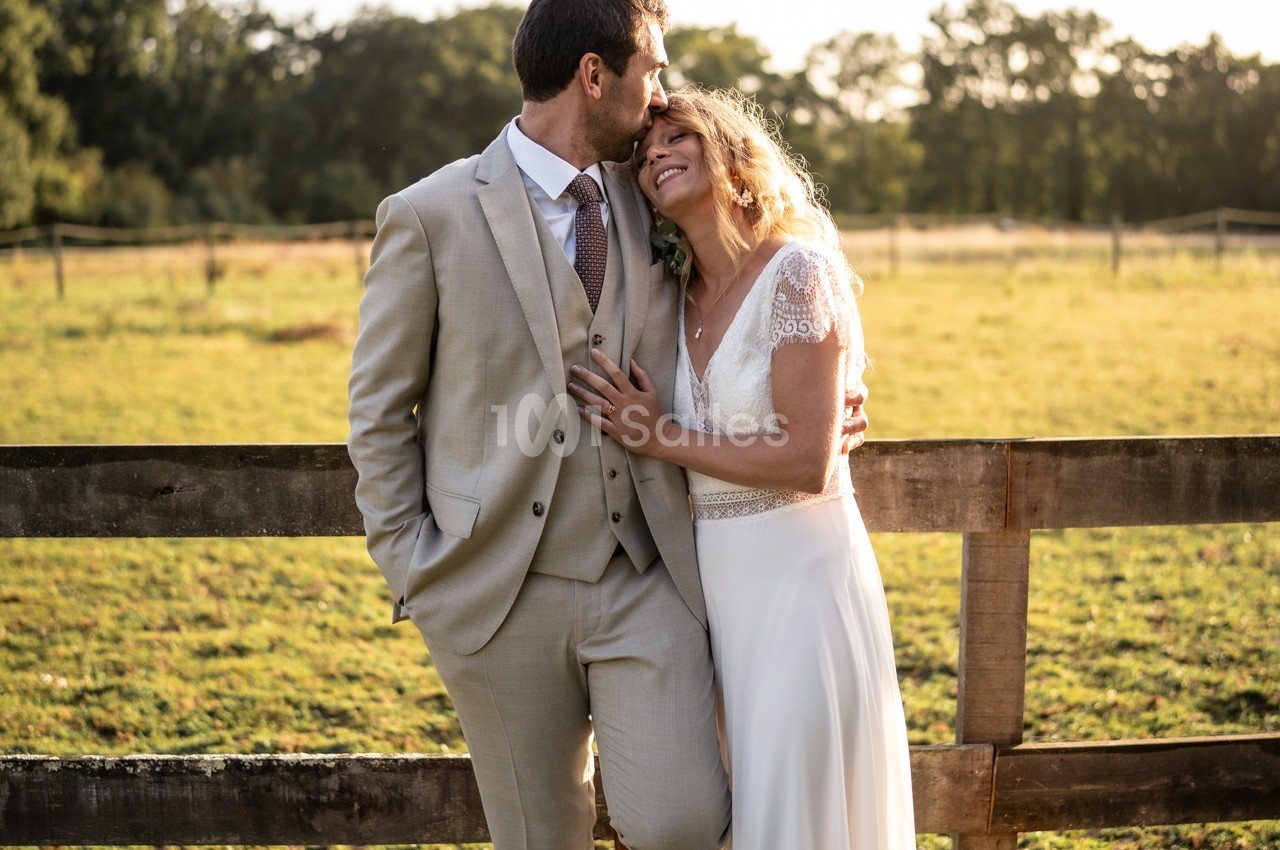 Un couple en tenue de mariage s'embrasse et sourit devant une clôture en bois dans un champ ensoleillé.