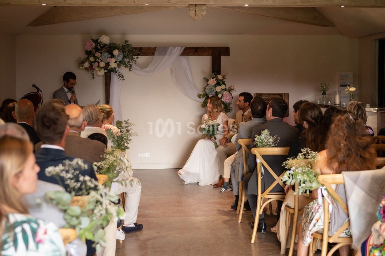 Cérémonie de mariage en intérieur avec des invités assis, un couple au centre et un officiant en train de parler.