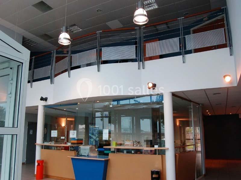 Accueil d'un bâtiment moderne avec comptoir en bois, éclairage suspendu et mezzanine avec garde-corps métallique.