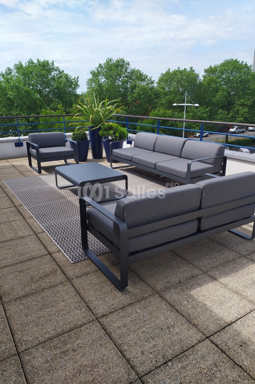 Salon de jardin moderne avec canapés gris, table basse et plantes, installé sur une terrasse ensoleillée avec vue arborée.