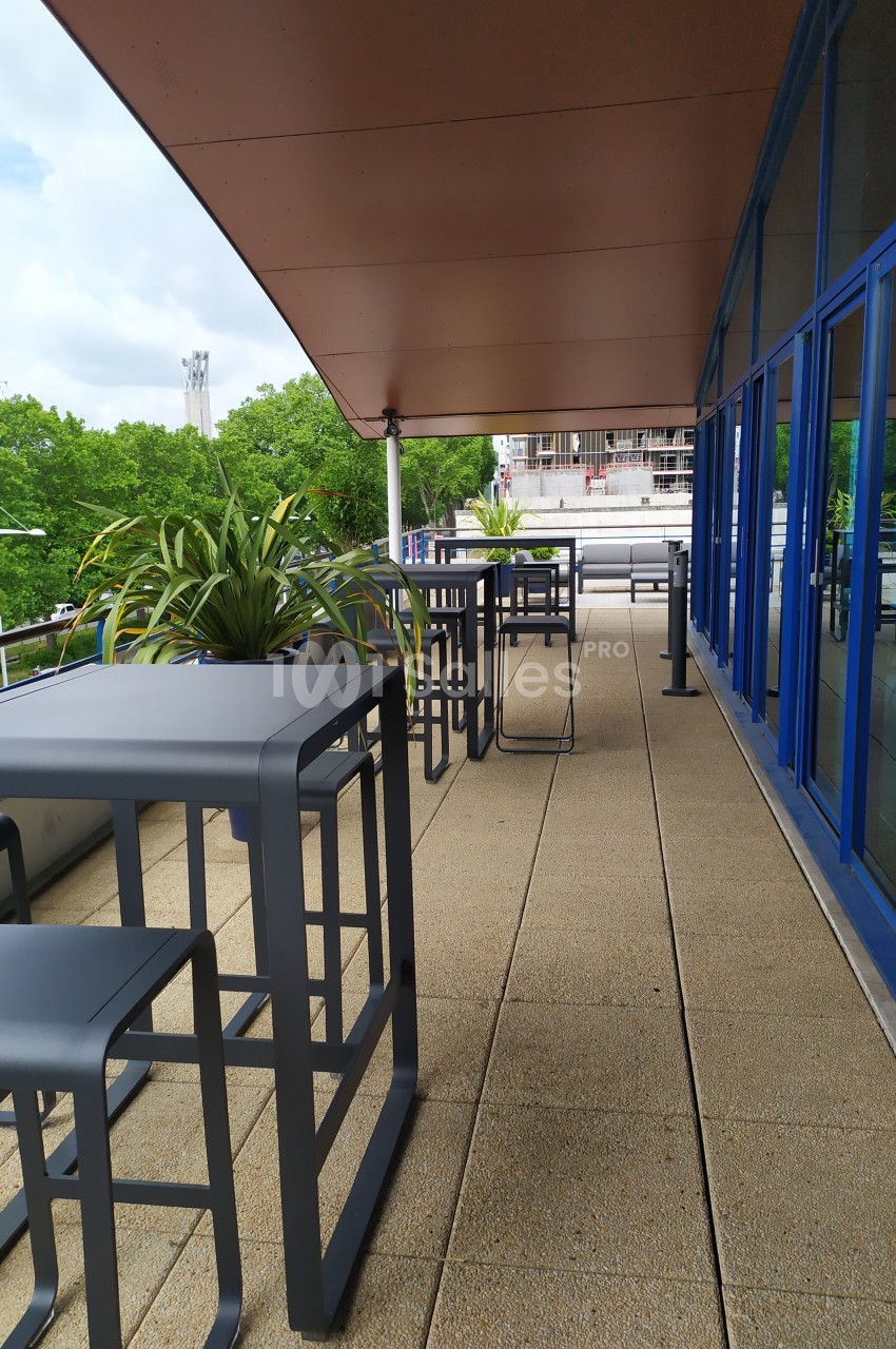 Terrasse avec tables et tabourets hauts en métal, entourée de plantes, donnant sur un paysage urbain et arboré.