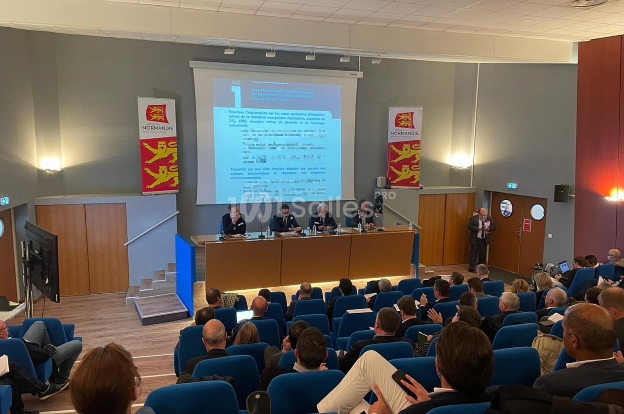 Salle de conférence avec des intervenants assis à une table et un public attentif dans des sièges bleus.