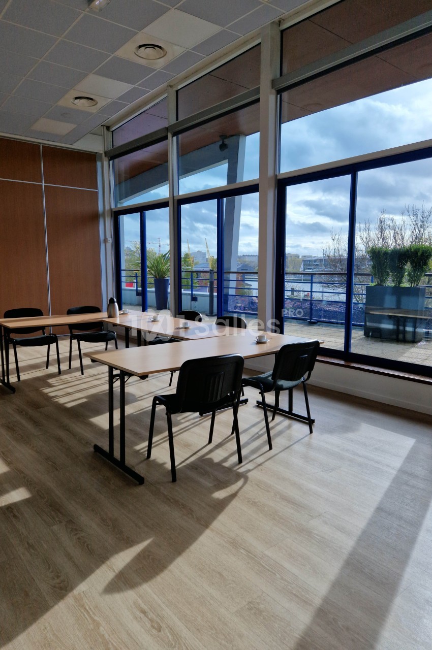 Salle de réunion lumineuse avec grandes baies vitrées, tables en bois, chaises noires et vue sur une terrasse arborée.