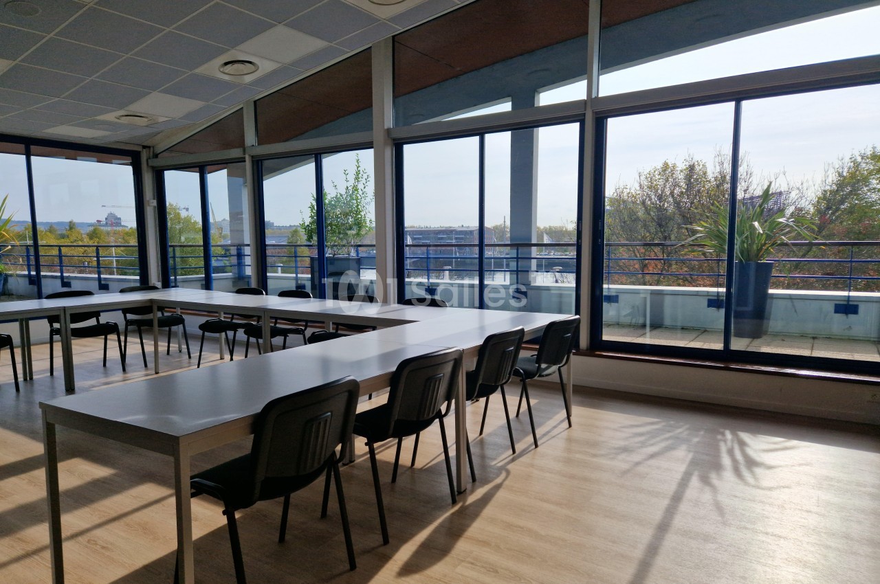 Salle lumineuse avec de grandes baies vitrées, des tables alignées et des chaises, donnant sur une terrasse avec des plantes.