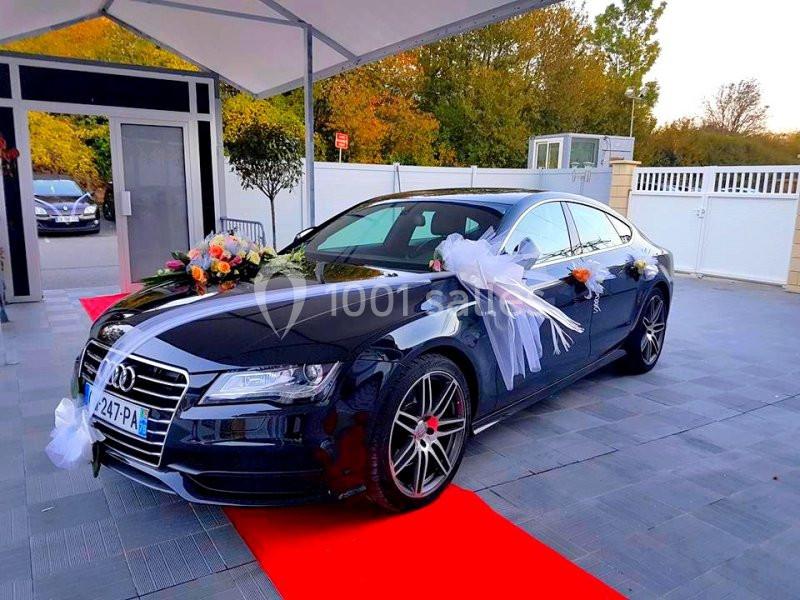 Voiture noire décorée de fleurs et rubans blancs, garée sur un tapis rouge devant un bâtiment moderne.