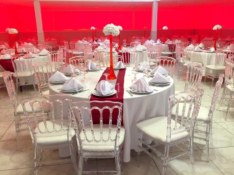 Salle de réception avec tables rondes dressées, nappes blanches, chemins de table rouges et chaises transparentes.