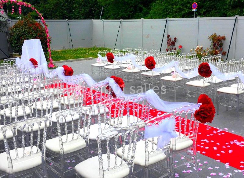 Allée centrale décorée de pétales roses et de rubans blancs, entourée de chaises transparentes avec fleurs rouges.