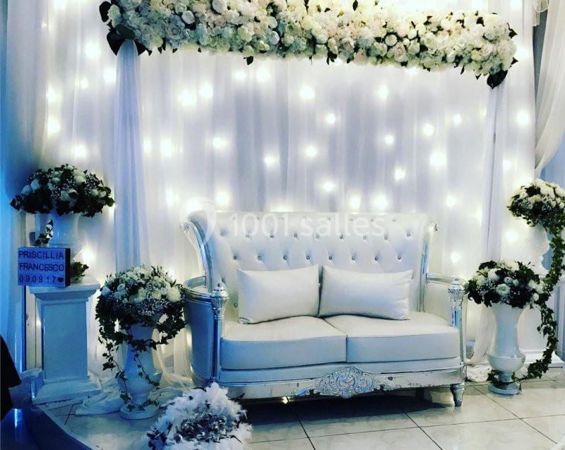 Canapé blanc capitonné devant un rideau lumineux décoré de fleurs blanches, entouré de compositions florales.