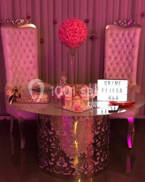 Table décorée avec des fleurs roses, accessoires élégants et deux chaises capitonnées blanches dans un cadre festif.