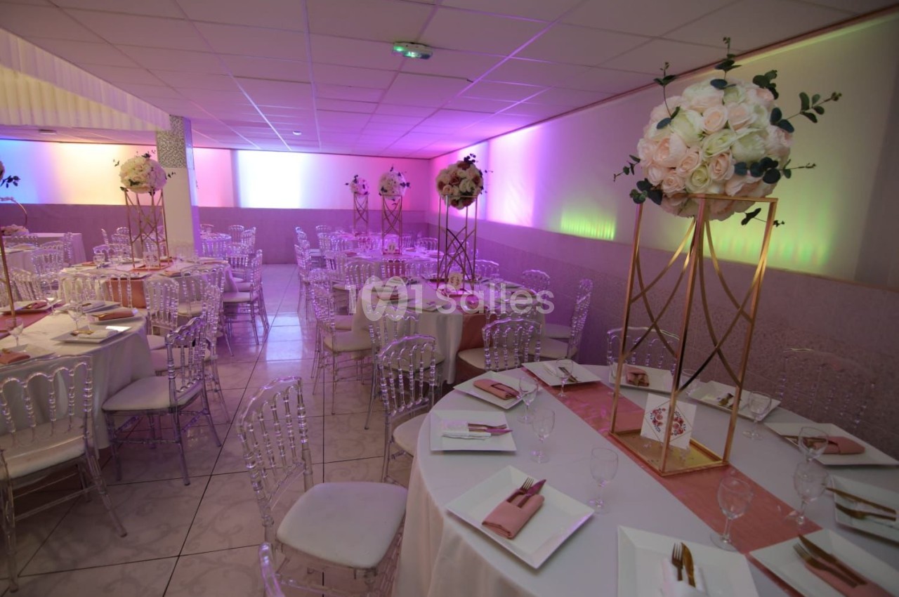Salle de réception décorée avec des tables rondes, nappes blanches, chaises transparentes et centres de table fleuris.