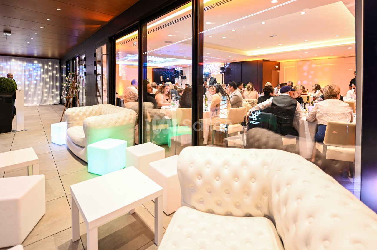 Salle de réception avec des invités assis à l'intérieur, vue depuis une terrasse avec fauteuils et tables lumineuses.