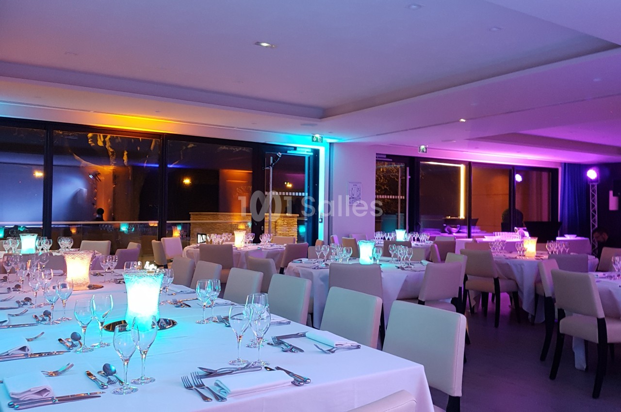 Salle de réception élégante avec tables dressées, nappes blanches, verres et lumières colorées en ambiance tamisée.