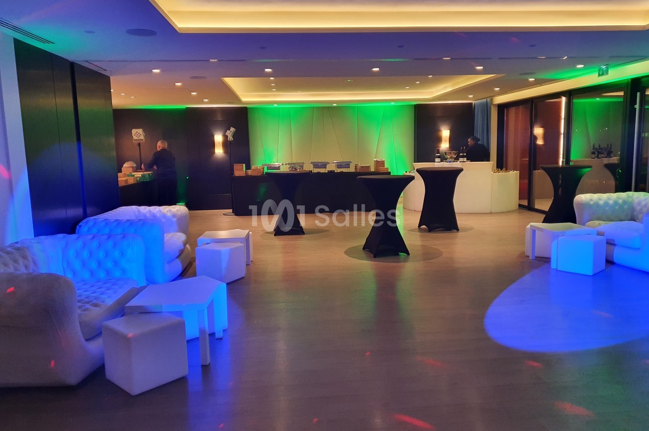 Salle de réception moderne avec éclairage coloré, tables hautes noires et canapés blancs disposés autour.