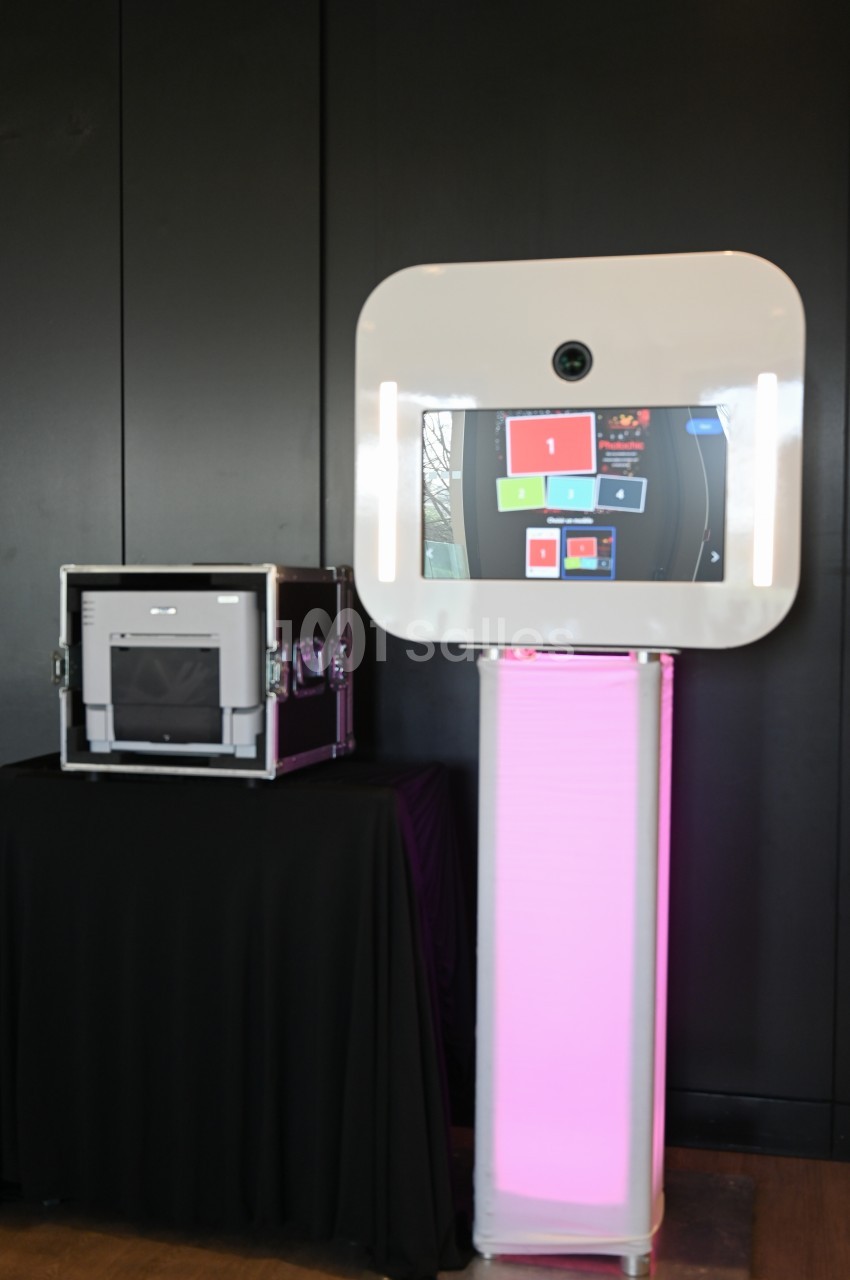Borne photo interactive sur pied avec écran lumineux, placée à côté d'une mallette noire dans un environnement intérieur.