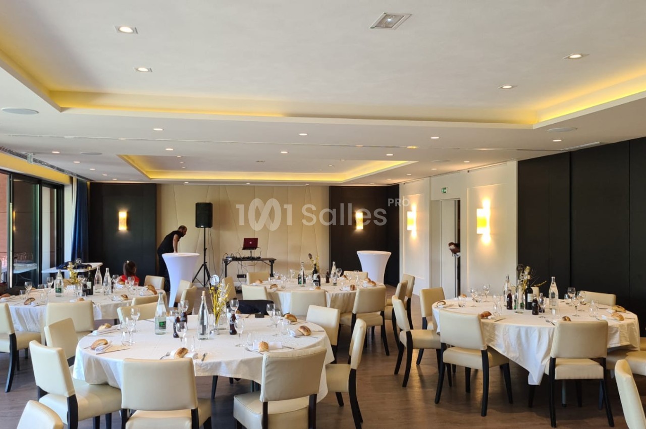 Salle de réception lumineuse avec tables rondes dressées, chaises beige clair et décoration sobre.