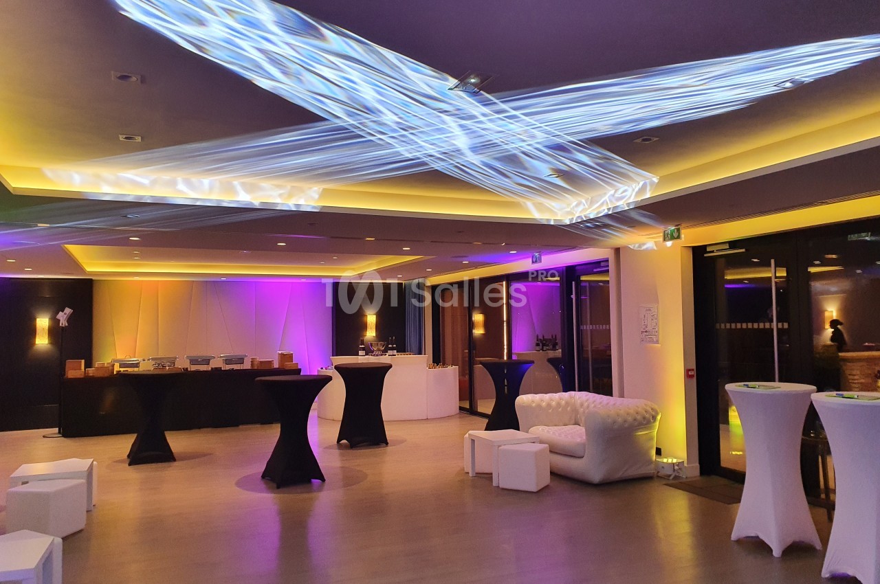 Salle de réception moderne avec éclairage coloré, tables hautes, fauteuil blanc et buffet en arrière-plan.