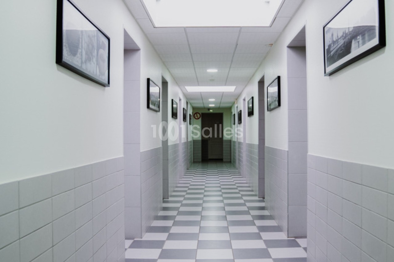 Couloir intérieur avec carrelage à motifs en damier, murs blancs ornés de cadres noirs et éclairage au plafond.
