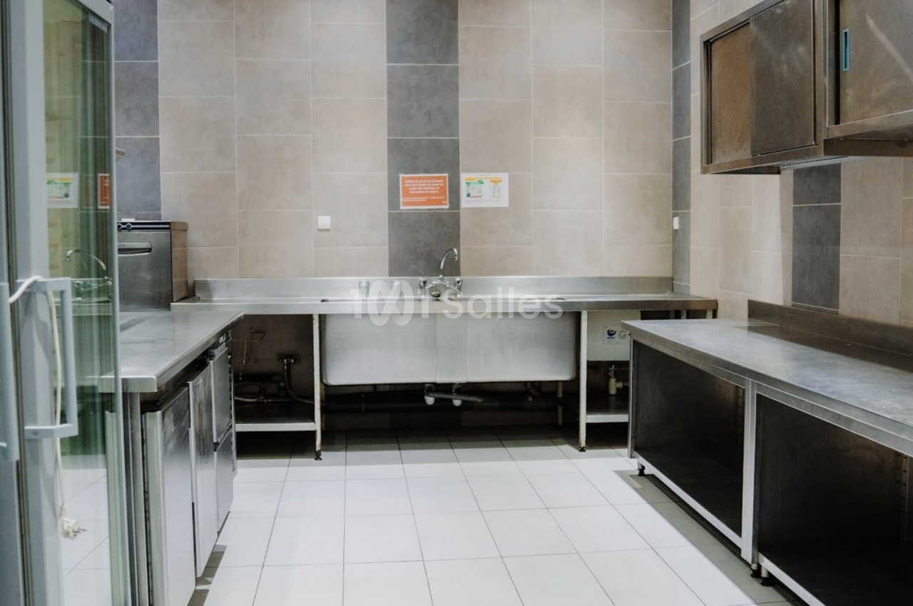 Cuisine professionnelle avec plans de travail en inox, évier central, réfrigérateur vitré et murs carrelés.