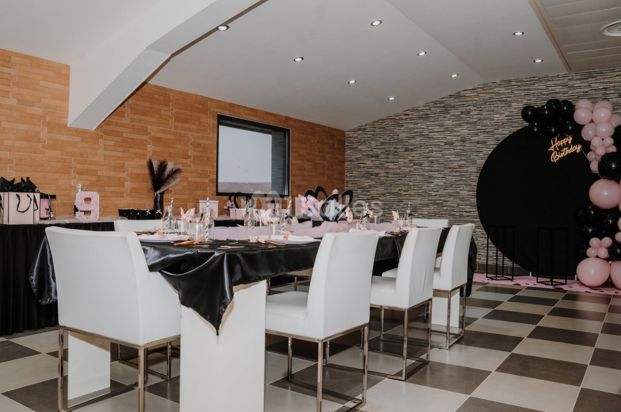Salle décorée pour une fête avec table dressée, ballons roses et noirs, et cadeaux sur une table latérale.