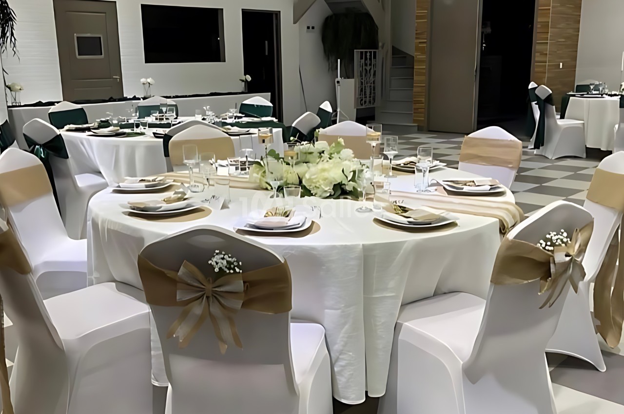 Salle de réception avec tables rondes dressées, chaises décorées de nœuds dorés et ambiance élégante.