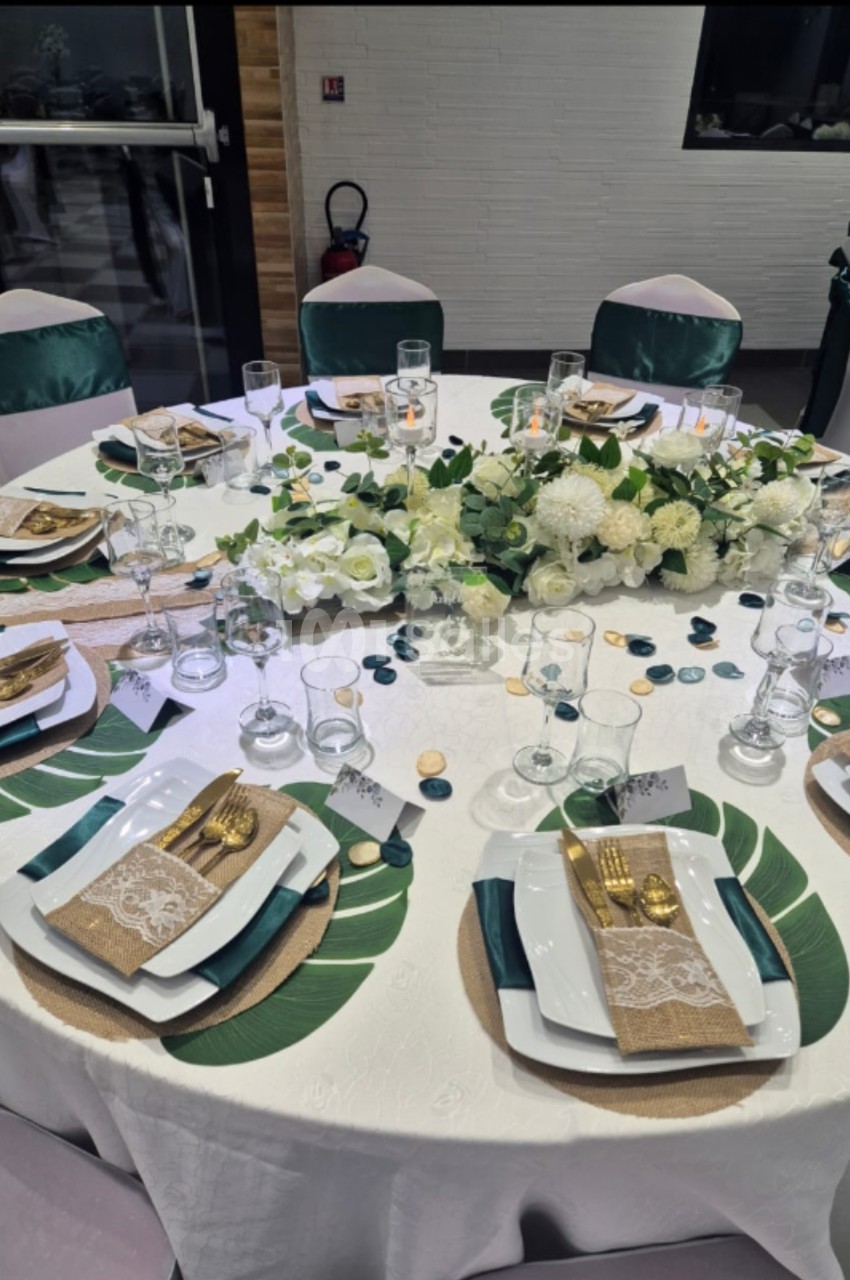 Table ronde élégamment dressée avec vaisselle blanche, couverts dorés, décorations florales blanches et feuillages verts.